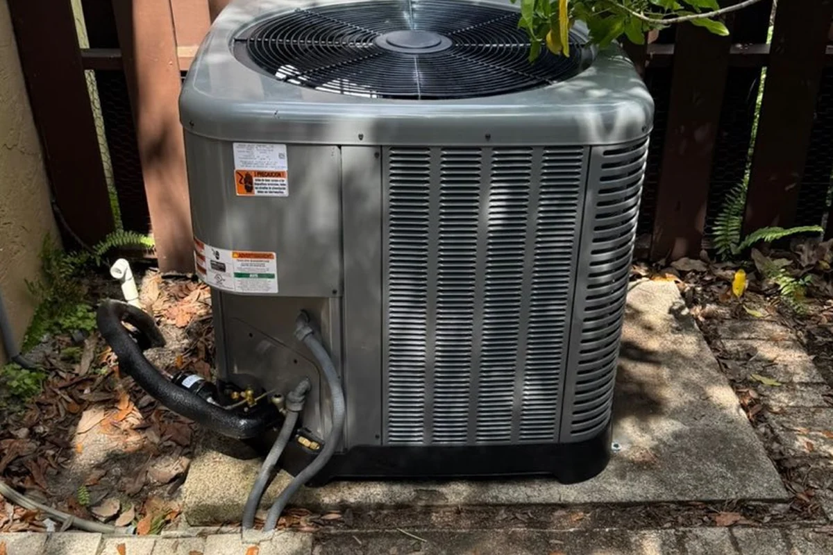 Outdoor air conditioning condensing unit beside a house, with the fan grille on top and coil fins around the cabinet