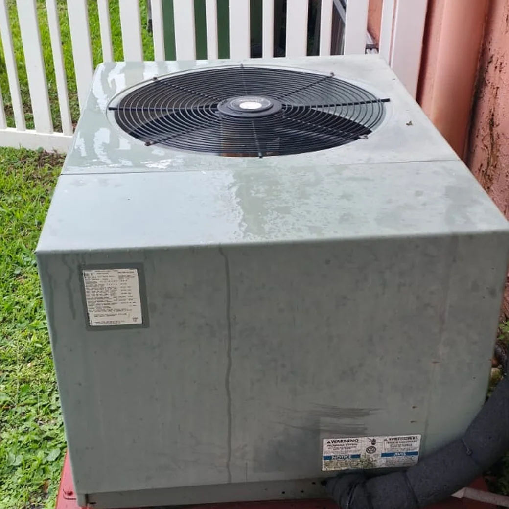 Round fan grille on top of a residential air conditioner condenser cabinet beside a house