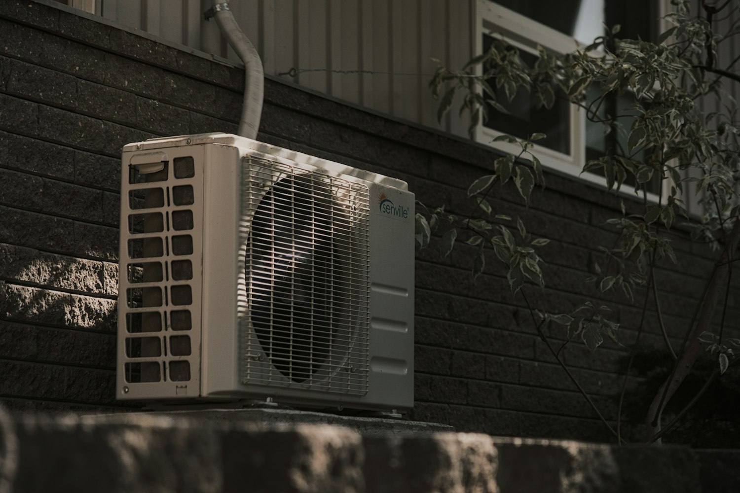 Residential outdoor AC condenser unit on a home exterior, with the condensate line running down the side wall